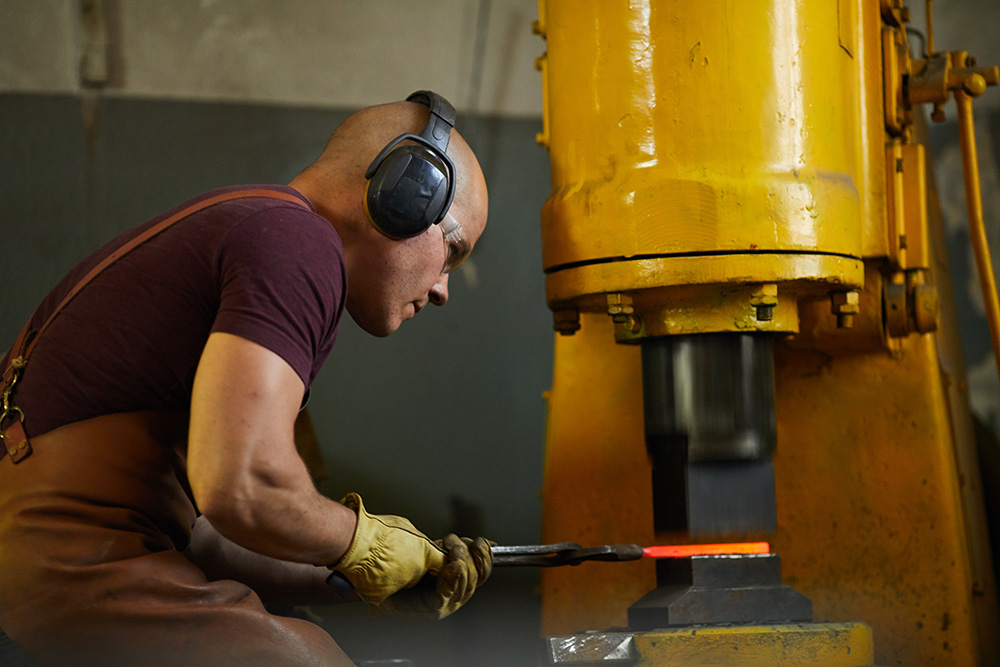 Startime Services: Arbeiter mit Gehörschutz und Schutzausrüstung bearbeitet Metallteil an Maschine in der Metallproduktion.