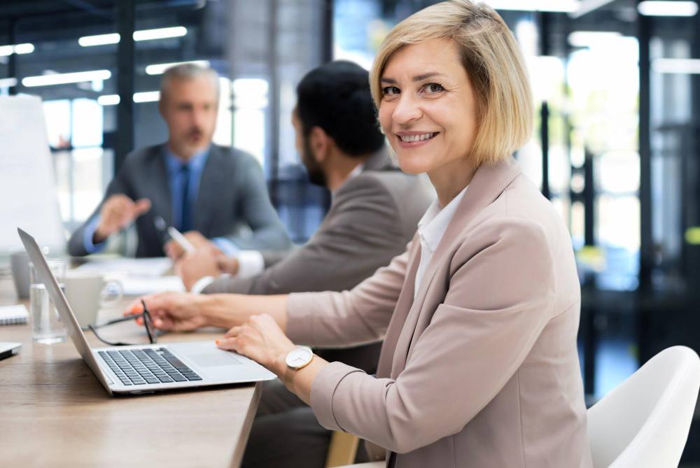 Geschäftsfrau im Büro mit Laptop und Kollegen im Hintergrund symbolisiert die Arbeitnehmerüberlassung von Startime Services für kurzfristigen und langfristigen Personalbedarf.